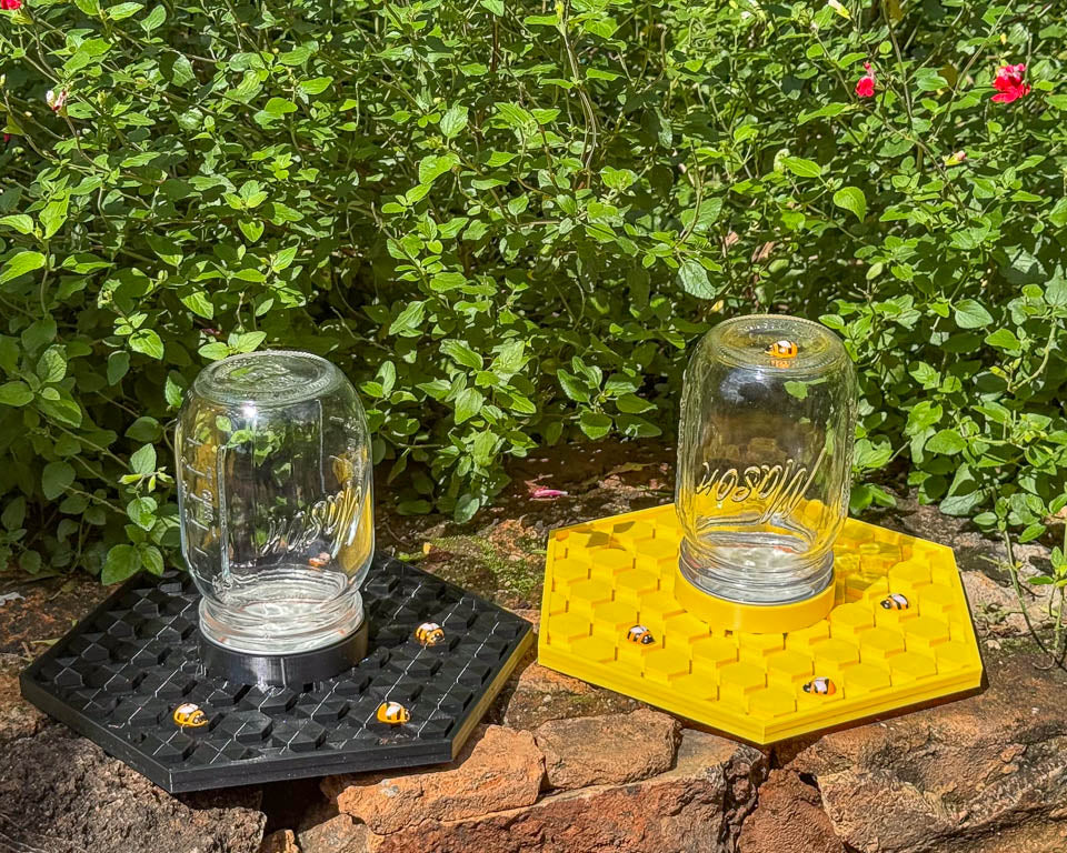 Two glass jars on hexagonal stands, one black and one yellow, in a garden with water for bees to drink safely
